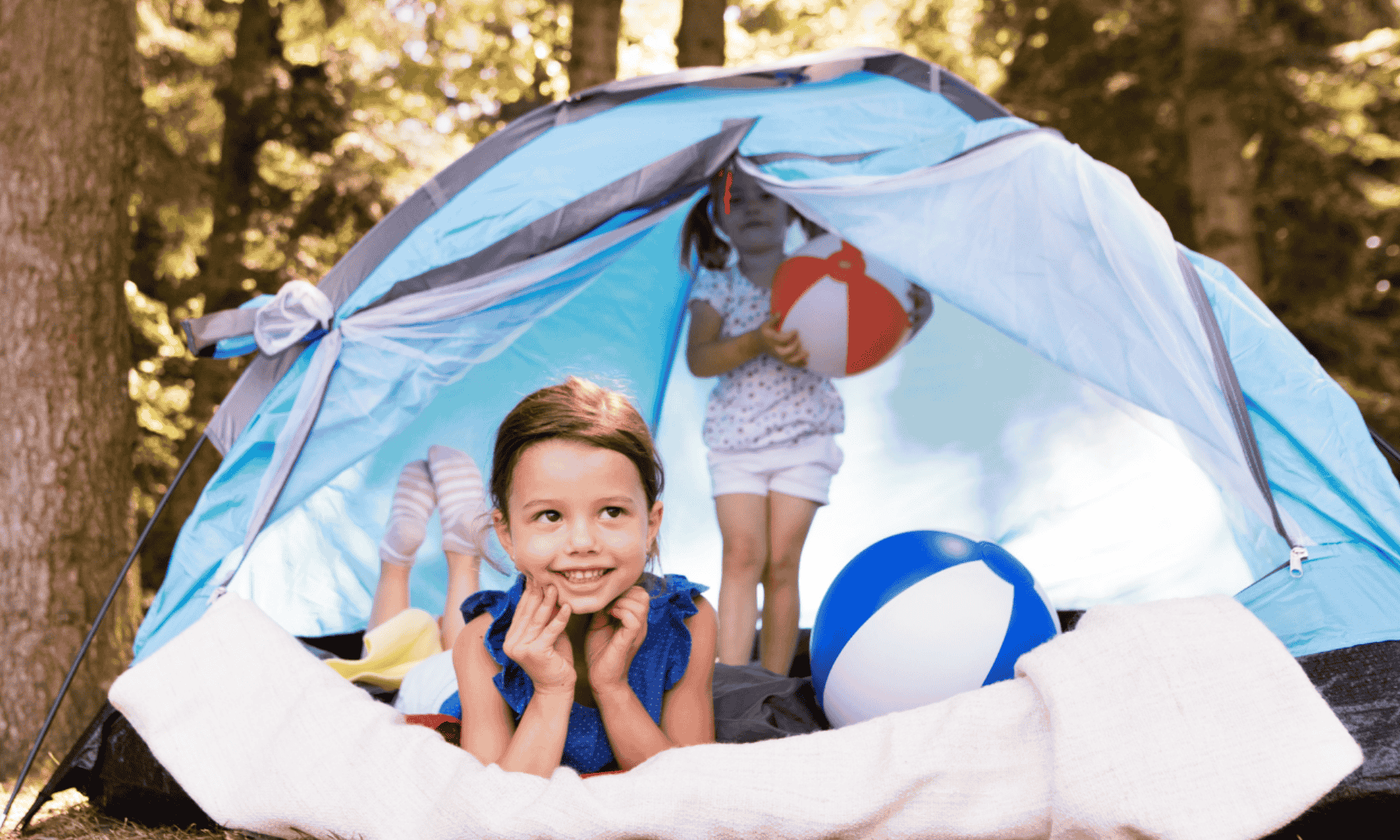Two children are inside a small outdoor tent, with blankets and beach balls placed on the ground in a wooded area.
