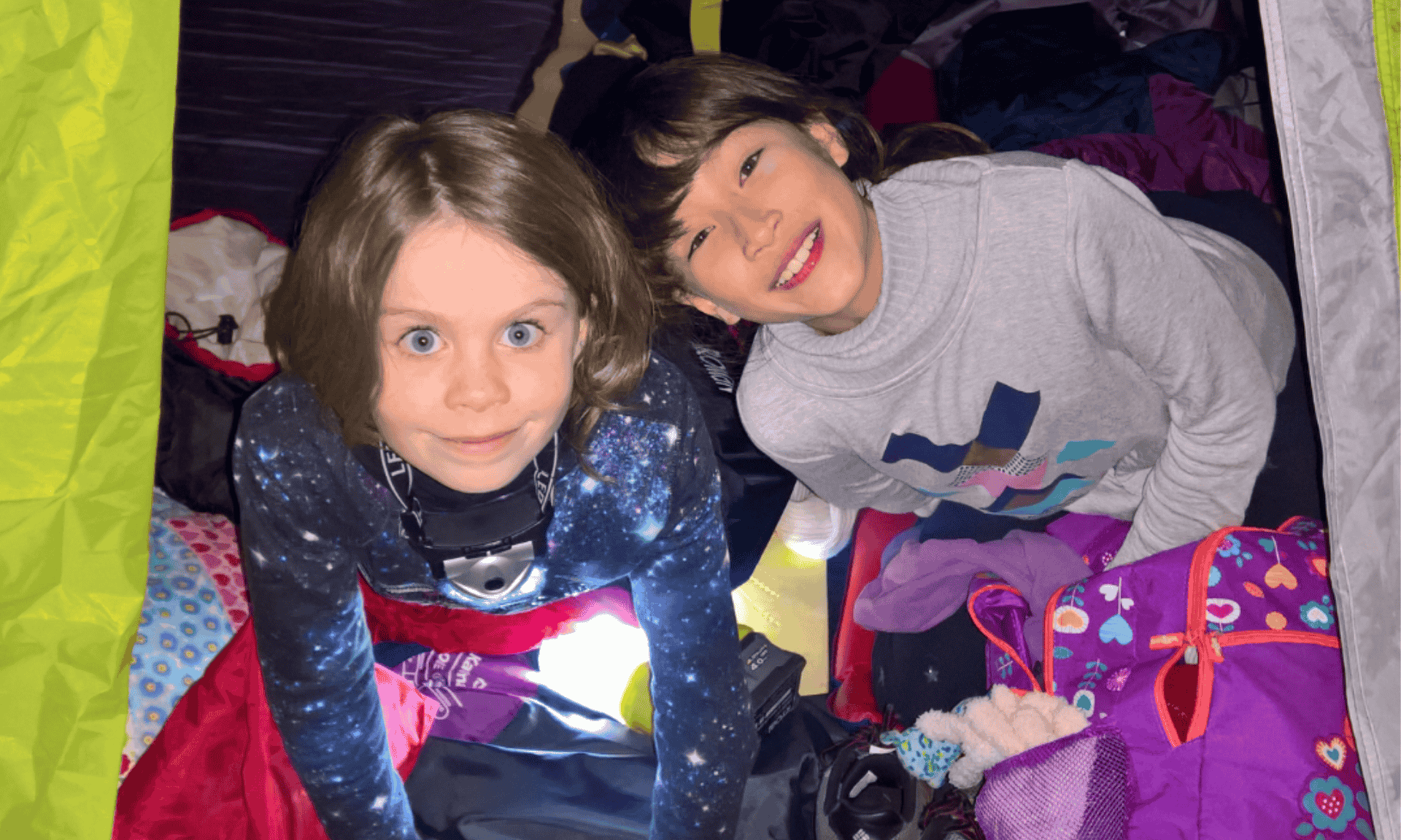 Two children sit inside a green camping tent with sleeping bags, clothing, and personal belongings arranged around them.