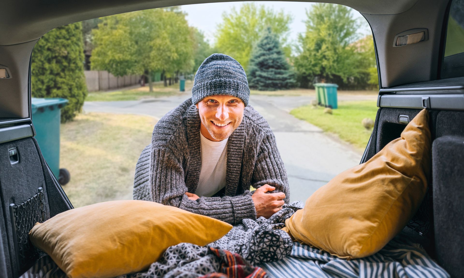 A person stands at the open back of a car with blankets and pillows arranged inside, parked on a quiet suburban street.