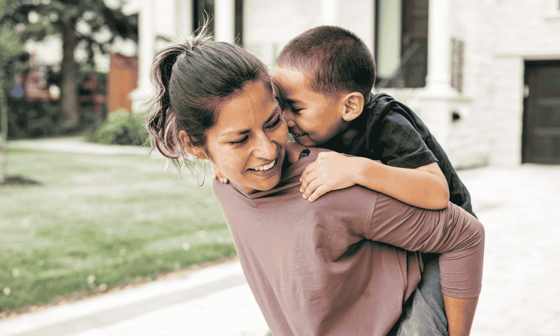 Smiling woman giving her young son a piggyback ride outside a modern home on a sunny day.